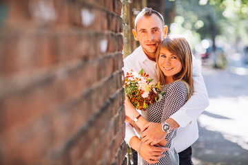 Fototapeta premium Close up fashion romantic portrait of young pretty couple kissing and hugs on the street at evening bright sunlight. Blonde gril with flowers boquet