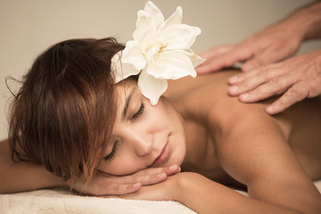 Bellissima ragazza con fiore in testa riceve un massaggio su un lettino di una spa, mentre si...