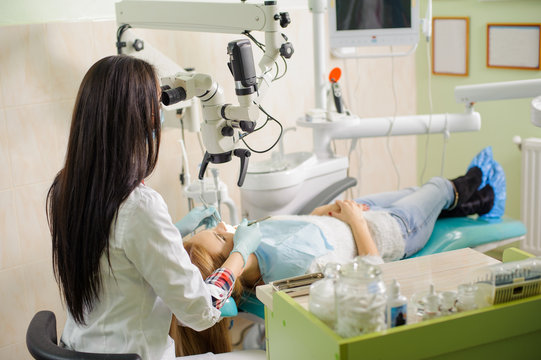 Young beautiful blonde woman petient lying on dentist chair with open mouth. Female dentist doctor treating root canals using microscope at dentistry office. Dental care and treatment.