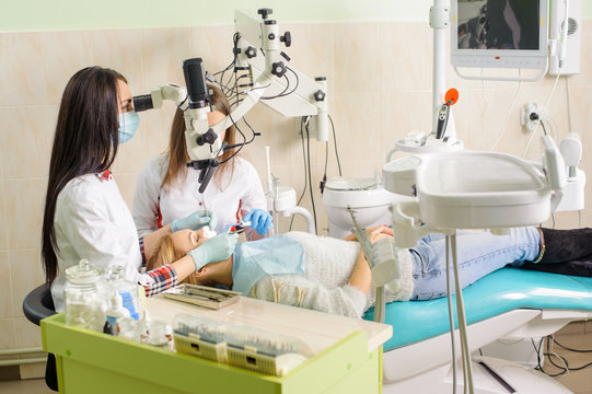Female dentist and her assistant treating caries using microscope, wearing masks and gloves. Young blonde woman petient lying on dentist chair. Medical equipment. Dental clinic