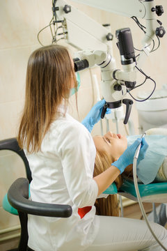 Female dentist doctor treating root canals using microscope at dentistry office. Young beautiful blonde woman petient lying on dentist chair with open mouth. Dental care and treatment.