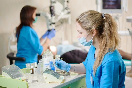 Young female dentist assistant taking dental drill on foreground. Doctor treating caries using microscope in the background. Dental equipment