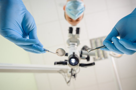 Male dentist is holding dental tools and working with a microscope in a dental clinic. First-person view from below. Focus on the tools. Oral procedure concept. Stomatology equipment