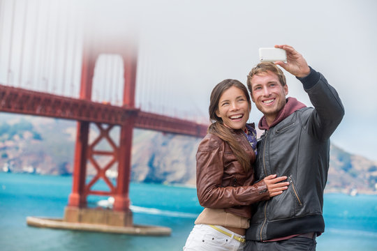Couple tourists taking selfie photo in San Francisco by Golden Gate Bridge. Interracial young modern couple using smart phone by famous american landmark. Asian woman, Caucasian man.