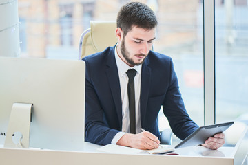 Businessman sitting behind a table holding a tablet and writes notebook