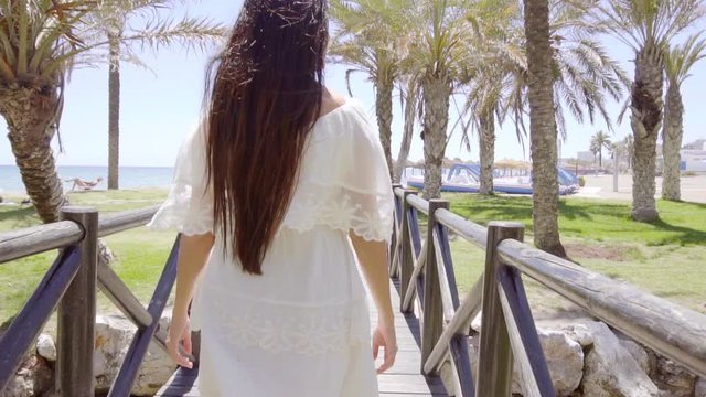 Young pretty smiling woman in white dress looking back while walking to beach with palm trees and green grass