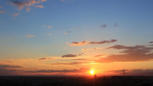 Sunset time-lapse with orange sun, blue sky, orange, yellow, grey clouds in the silhouette of the town