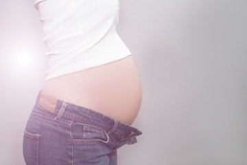 Pregnant girl in unbuttoned jeans and white t-shirt holding on to naked belly. The studio with gray background