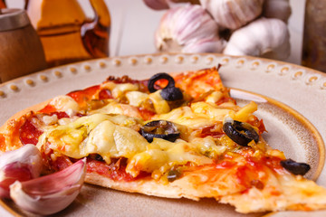Homemade pizza out of the oven with ham, olives, tomato and cheese on baking paper. On the table in light wood. Pepper mill and spices, whole garlic.