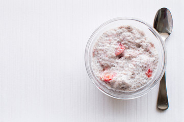 High angle view of chia seed and strawberry pudding in glass dish with spoon on white textured background