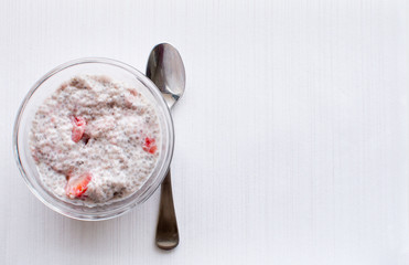 High angle view of chia seed and strawberry pudding in glass dish with spoon on white textured background