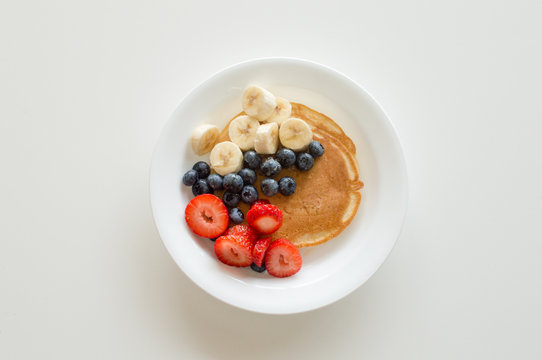 High angle view of pancake with banana, blueberries, strawberries and honey on white plate against white background