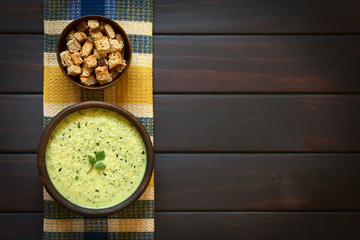 Cream of zucchini soup garnished with parsley leaf, with a bowl of homemade croutons on kitchen towel, photographed on dark wood with natural light