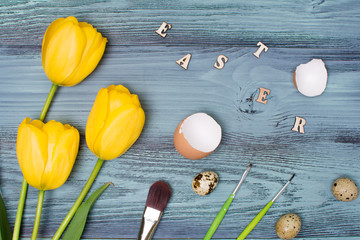 Easter eggs, tulips and brushes over blue wooden table. Top view.