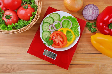 Plate with sliced fresh vegetables on digital kitchen scales over wooden background