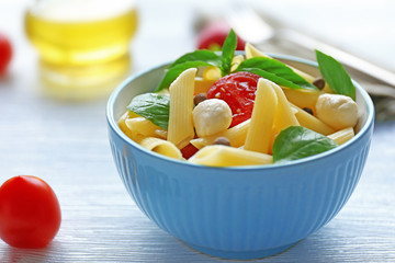 Cooked penne pasta with mozzarella, fresh tomatoes and basil in blue bowl on wooden table