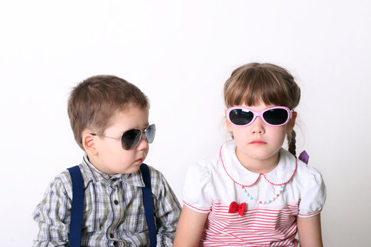 Little serious boy and girl sit in sunglasses in white studio
