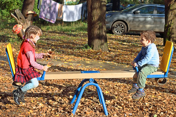 Girl in red vest and her brother ride on seesaw at sunny fall