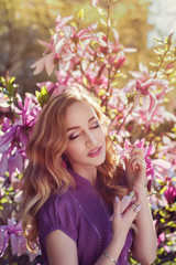 Fototapeta premium Outdoor portrait of a young beautiful lady using perfume near magnolia tree with flowers. Model closed her eyes. Girl wearing stylish clothes. Female spring fashion