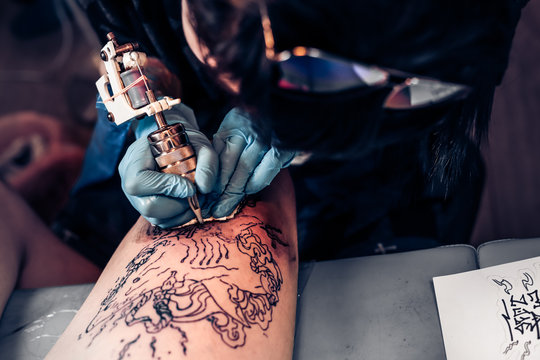 Closeup tattoo process in a professional salon. Master makes tattoo pattern on the client leg. Master works in sterile blue gloves.