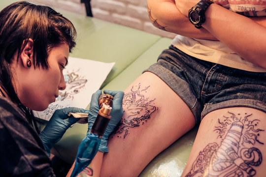 Professional tattoo artist doing tattoo. Portrait of a woman tattoo master showing a process of creation tattoo on a legs. Tattooist machine,gun. Free space to fill,text.