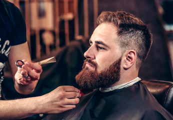 Closeup professional grooming beard with scissors in a Barbershop. Portrait of a bearded man hipster in a Barbershop during haircut. Stylish man hair cut at the barber shop.  