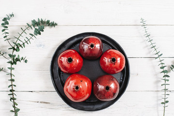 Many ceramic red pomegranates and black plate on white wooden background. Flat lay set of color  house decor. Organic food, pottery, decoration concept. Bright ceramic red pomegranates on black dish