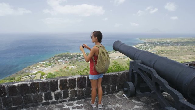 Tourist visiting St. Kitts Brimstone Hill Fortress National Park on vacation on St Kitts and Nevis. Caribbean cruise ship destination. Girl standing taking photo using smart phone camera by cannon.
