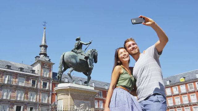 Tourists couple taking selfie using smart phone in Madrid Spain on Plaza Mayor. Happy people taking photos. Young woman and man on famous square in front of statue of Felipe 3rd. RED EPIC SLOW MOTION.