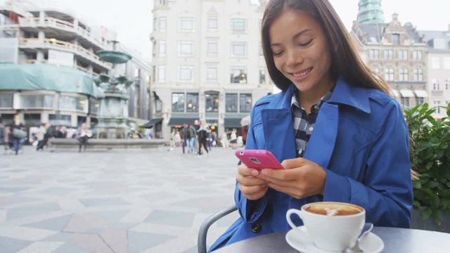 Asian business woman texting or reading news on smart phone drinking coffee at outdoor terrace cafe. Stroget, Copenhagen, Denmark, Europe. SLOW MOTION, RED EPIC.