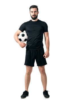 Smiling soccer or futsal player wearing black sportswear holding ball under his arm looking at camera. Full body length portrait isolated over white background.