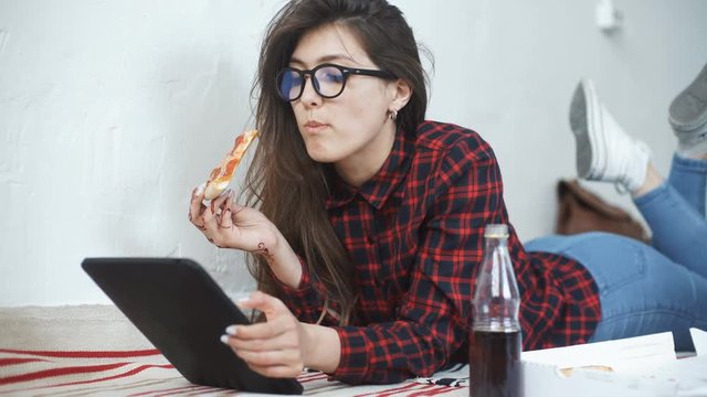 Asian woman eating pizza and looking at digital tablet computer at home