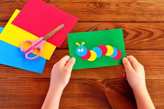 Children hands holding paper applique caterpillar. Paper sheets, scissors on a brown wooden background