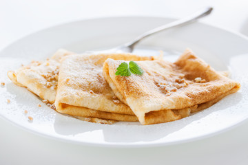 French crepes or pancakes with sugar powder, nuts and fresh mint on a white plate on a white background for breakfast, closeup