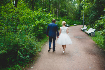 Fototapeta premium A man in a suit and a woman in a wedding dress walking in the Park in the summer. Short wedding dress