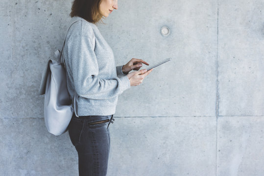 Young smiling hipster firl using digital tablet outdoors, concrete wall on the background with copy space for your text message or desing