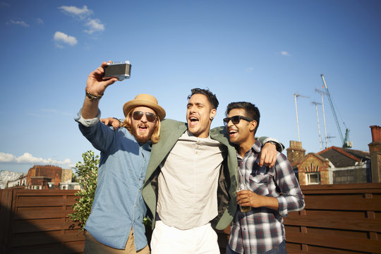 Three male friends taking camera selfie at rooftop party