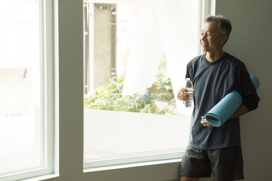 Mature man holding rolled up yoga mat and bottle of water looking out of window