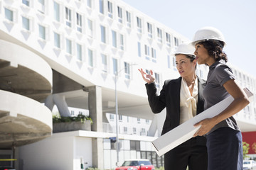 Two businesswomen, wearing hard hats, carrying plans