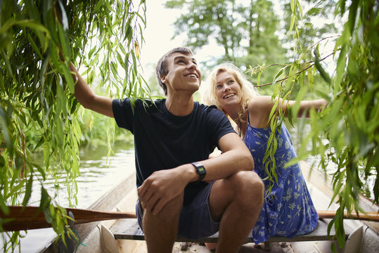 Young couple touching willow tree foliage from rowing boat on rural river