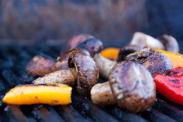 Seasoned mushrooms and peppers are being grilled on iron grates over charcoal