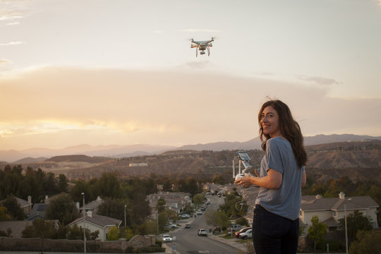 Female commercial operator flying drone above housing development, looking over shoulder at camera, Santa Clarita, California, USA