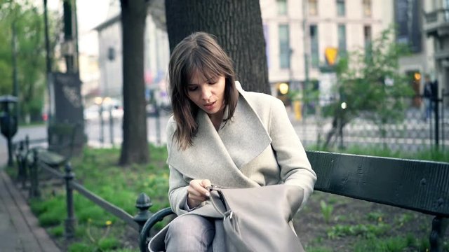 Young woman searching for her phone in handbag sitting on bench in park
