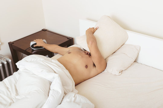 Young man in bed, covering face with pillow, turning off alarm clock