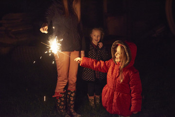Side view of girl wearing red fur trim coat holding sparkler being ignited by mother, smiling