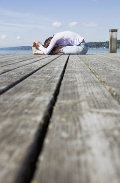 Surface Level Side View Of Mature Woman Sitting On Pier Bending Over Touching Toes