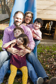 Family in a row between each others legs on playground slide, looking at camera smiling