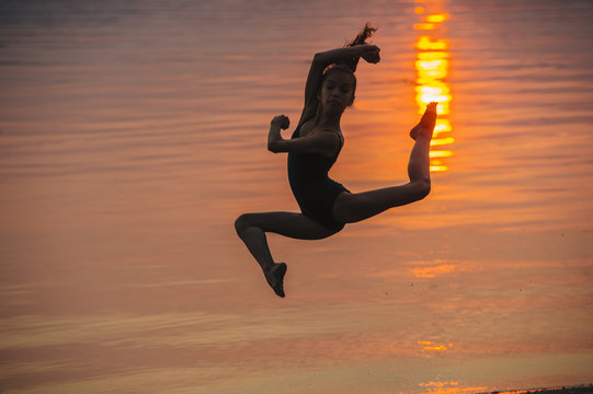 Side view of girl in silhouette by ocean at sunset leaping in mid air, legs apart looking at camera
