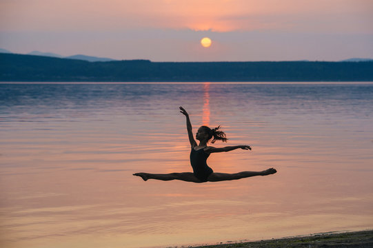 Side view of girl by ocean at sunset leaping in mid air, arms raised doing the splits