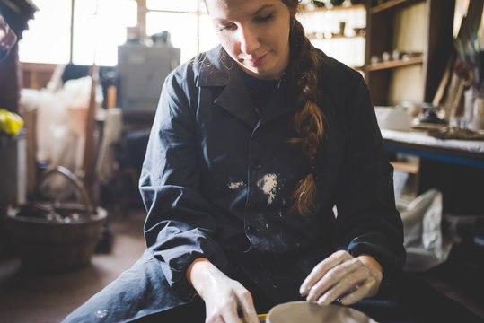 Front view of young woman sitting at pottery wheel looking down making clay pot - Powered by Adobe